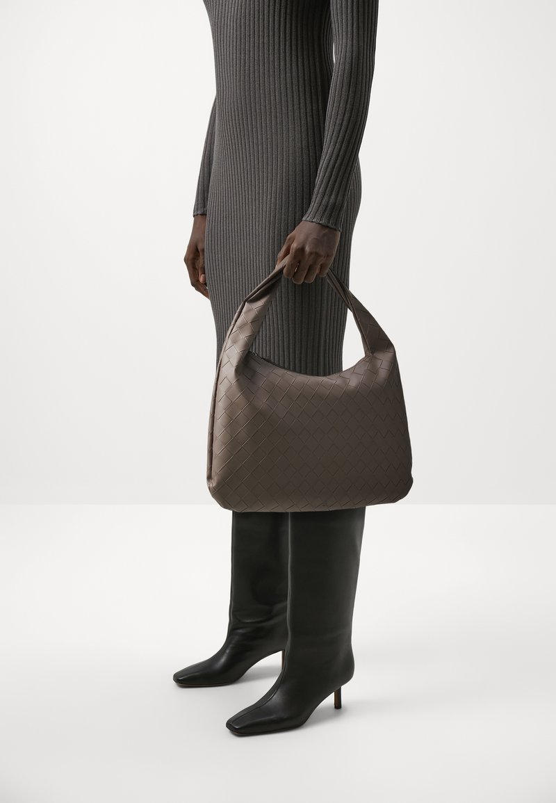 Brązowa, pleciona torebka ze skóry z wyraźnie zarysowanym kształtem, z okrągłym uchwytem. Modelka nosi szarą sukienkę o prążkowanej fakturze oraz czarne kozaki do kolan.