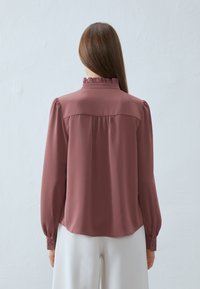 Blusa con colletto arruffato in rosa polveroso, realizzata in tessuto morbido. Presenta maniche lunghe con polsini abbottonati e una piega sul retro per un maggiore movimento.