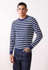 Marineblauwe en witte gestreepte trui van gebreide stof, met een ronde hals en een klein rood logo op de borst. Draag het met donkere jeans.
