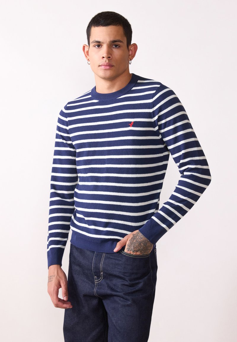 Marineblauwe en witte gestreepte trui van gebreide stof, met een ronde hals en een klein rood logo op de borst. Draag het met donkere jeans.