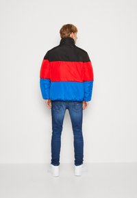 Giacca imbottita con una parte superiore nera, un centro rosso e una parte inferiore blu. Indossata sopra jeans scuri e sneakers bianche, presenta un colletto alto.