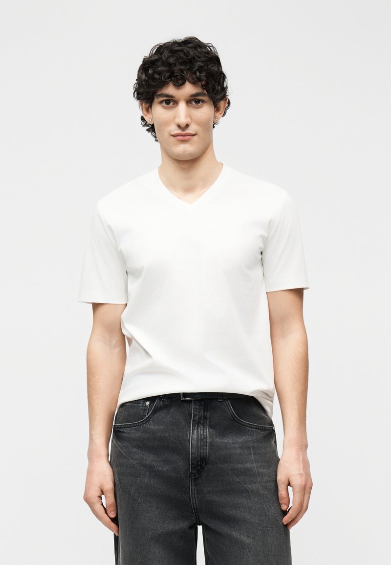 Νεαρός με σγουρά σκούρα μαλλιά φοράει λευκό μπλουζάκι με V λαιμόκοψη και κοντά μανίκια, καθώς και μαύρο τζιν, στέκεται μπροστά σε λευκό φωτεινό φόντο.