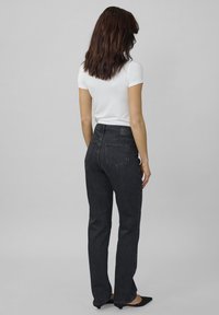 Frau mit schulterlangem braunem Haar, die ein weißes Kurzarmoberteil, schwarze hoch taillierte Jeans und schwarze spitze Ballerinas trägt, steht mit dem Rücken zur Kamera.