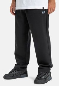 Czarne dresy o luźnym kroju, wyposażone w boczne kieszenie i małe logo marki na lewej udzie, w połączeniu z czarnymi butami sportowymi.