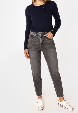 Kobieta ubrana w granatową bluzkę z długim rękawem, szare, wyblakłe dżinsy z wysokim stanem oraz białe trampki, stoi z jedną ręką w kieszeni.