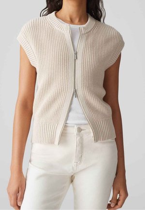 Femme portant un gilet en maille beige sans manches avec une fermeture éclair sur le devant, par-dessus un haut blanc et un pantalon couleur crème, vue du cou aux hanches.