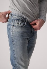 Světlé modré džíny vyrobené z denimu s vybledlým vzhledem, s pěti kapsami a kovovým zapínáním. Látka vypadá mírně opotřebovaně.