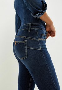 Tmavě modré skinny džíny s hladkou texturou, mají středně vysoký pas, dvě zadní kapsy a kontrastní stehy.