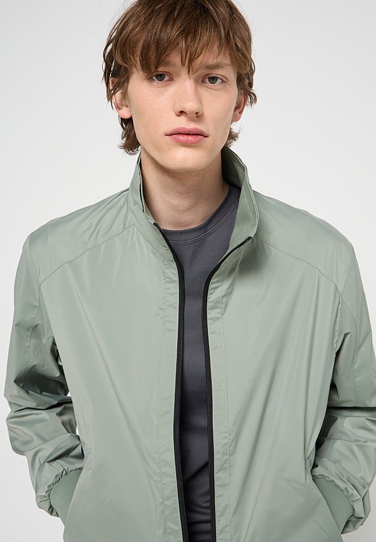 Herren Jacke in Hellgrün, aus glattem, leichtem Material. Mit einem Frontreißverschluss, Stehkragen und lockerer Passform. Trägt man über einem dunklen Hemd.