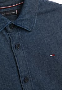 Denimová košile v tmavě modré barvě s límečkem na knoflíky, vybavená černými knoflíky a malým vyšitým logem v červené, bílé a modré barvě na hrudi.