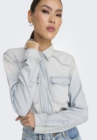 Mujer joven con el cabello recogido, vistiendo una camisa de mezclilla azul claro y jeans, posando con una mano cerca del mentón y una expresión neutral.