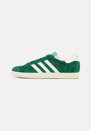 Groene suède sneaker met witte leren strepen, rubberen zool, vetersluiting en contrasterend groen detail op de tong. Voorzien van Adidas-logo.