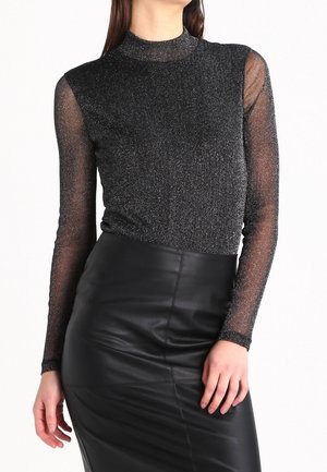 Femme portant un haut noir transparent pailleté à manches longues, rentré dans une jupe en cuir noire taille haute avec des détails de couture.