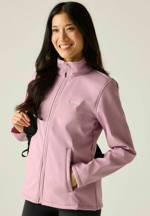 Femme aux longs cheveux foncés portant une veste rose clair zippée, souriante et tenant une sangle de sac à dos noire sur son épaule.