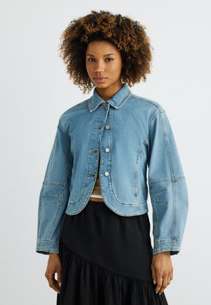 Göndör hajú nő világoskék, gombos farmerdzsekiben, fekete szoknyával, egyszínű háttér előtt.