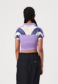 Polo cropped em roxo e branco, com mangas curtas, colarinho e padrões geométricos nas costas. Combinado com calções cargo pretos.