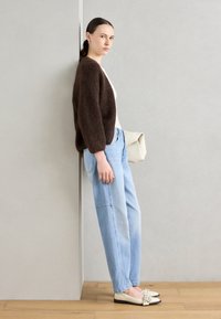Brauner, flauschiger Cardigan, weißes Oberteil, hellblaue, lockere Jeans, beigefarbene Schultertasche und cremefarbene Ballerinas mit schwarzem Boden und Schleifen-Detail.