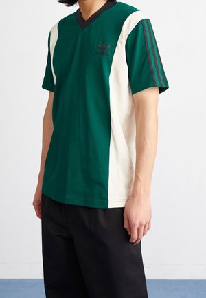 Grünes Baumwoll-T-Shirt mit cremefarbenen und schwarzen Akzenten, V-Ausschnitt-Design, mit dem Adidas-Logo und drei schwarzen Streifen an den Ärmeln.