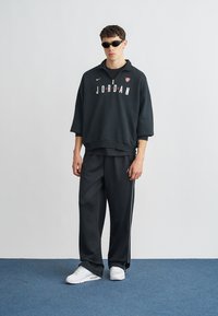 Μαύρο φούτερ με κουκούλα και λογότυπο "JORDAN", φαρδιά εφαρμογή, με rib μανσέτες· συνδυασμένο με μαύρο παντελόνι φόρμας με πλευρικές ρίγες και λευκά αθλητικά παπούτσια.