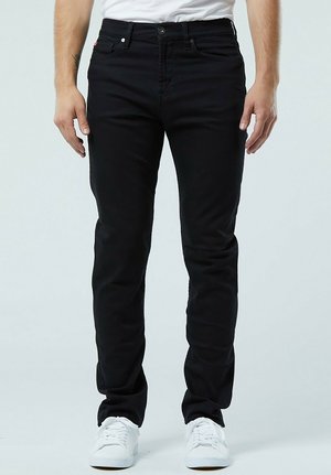 Jean slim noir en denim, avec cinq poches et une fermeture à bouton sur le devant. Associé à des baskets blanches.