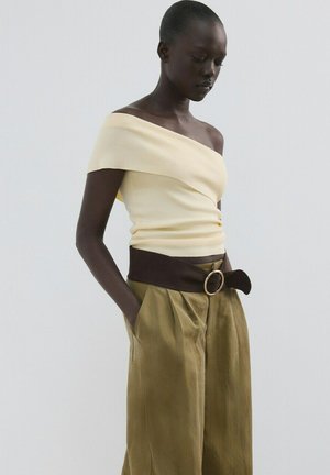 Kvinde iført en lys gul off-shoulder top og højtaljede olivengrønne bukser med et bredt mørkt bælte, stående med hænderne i lommerne.
