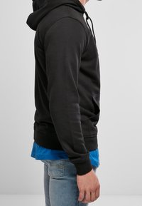 Czarna bluza z kapturem, z kieszenią kangurką, sznurkiem w kapturze oraz ściągaczami przy rękawach i u dołu. Noszona na niebieską koszulę, z jasnoniebieskimi dżinsami.