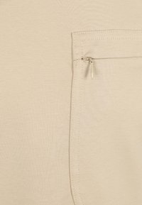 Beige stof met een gladde textuur heeft een klein ritszakje aan de rechterkant, benadrukt door nette stiksels langs de randen.