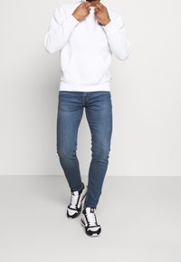 Bílá mikina s kapucí, modré slim fit džíny a černé, bílé a červené sportovní boty. Obsahuje přední kapsu a kontrastní akcenty na botách.