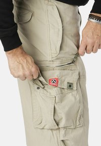 Lichte khaki cargobroek met meerdere zakken; een zichtbare rode "MOLECULE" label op de achterzak; materiaal lijkt duurzaam en gestructureerd.