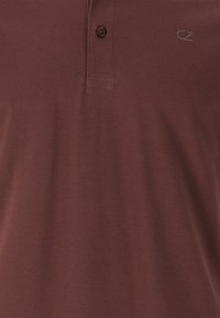 Brun polotröja med textilstruktur, två knappar vid kragen och en subtil "CZ"-logotyp broderad i en matchande nyans.