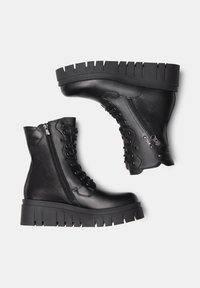 Bottes noires à la cheville avec une semelle en caoutchouc épaisse, un laçage à l'avant et une fermeture éclair sur le côté. Fabriquées en cuir lisse avec des détails de couture.