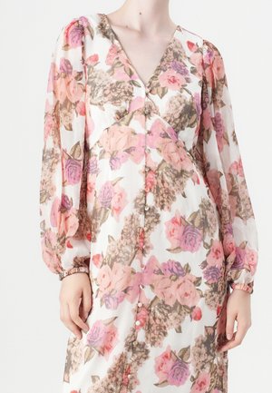 Frau trägt ein langärmliges Blumenkleid mit pinken, violetten und beigen Blumen und einem V-Ausschnitt mit Knöpfen vorne.