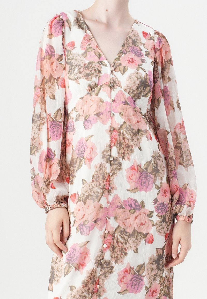 Femme portant une robe à manches longues à fleurs roses, violettes et beiges avec un décolleté en V et des boutons sur le devant.