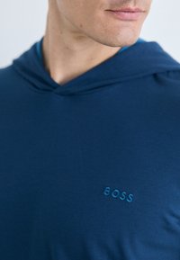 Marineblå hettegenser laget av mykt stoff med et brodert logo "BOSS" i matchende blå, med en avslappet passform og justerbar hals.