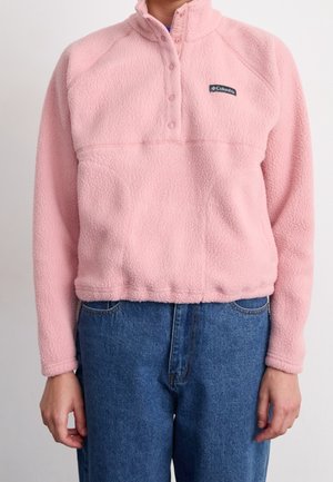 Henkilö, joka pukeutuu vaaleanpunaiseen Columbia-fleecepulloveriin, jossa on painonapit, ja sinisiin farkkuihin, seisoo yksiväristä taustaa vasten.