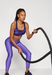 Violeta sportiskā krūštura virsdrēbe ar melnu joslu, apvienota ar spīdīgām violetām legingām. Models tur resnu melnu kaujas virvi, parādot aktīvu pozu.