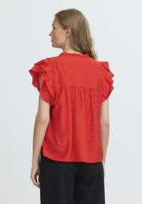 Camicetta rossa a maniche corte con spalle arricciate, tessuto strutturato e parte posteriore arricciata. Indossata con pantaloni neri. Scollo rotondo semplice.