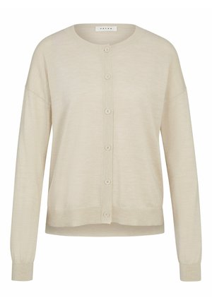 Beige Strickjacke mit Knöpfen, getragen über einem weißen Hemd und beigen Hosen. Das Modell steht in weißen Sneakers und präsentiert einen lässigen, geschichteten Look.