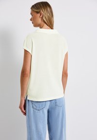 Donna con capelli biondi che sta di spalle, indossa una maglietta in maglia color crema a maniche corte e jeans di denim blu chiaro contro uno sfondo semplice.
