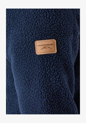 Donkerblauwe fleecejas met een gestructureerd oppervlak. Heeft een beige leren patch met het label "SWEDEMOUNT" en een bergafbeelding.
