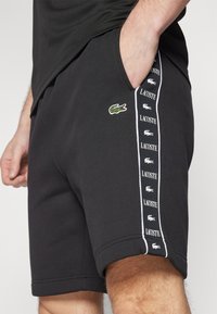 Črne kratke hlače iz bombaža; vsebujejo stranske žepe in izvezeno zeleno krokodilovo logotip. Bele črte z ponovljenim logotipom "LACOSTE" ob strani.