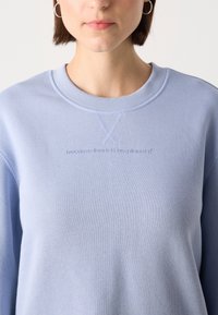 Jeune femme portant un sweat-shirt bleu clair avec un petit texte brodé "because there is no planet B" et des boucles d'oreilles anneaux argentées.