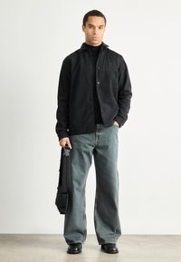 Casaco preto de botões, pulôver tipo gola alta por baixo, jeans de perna larga azul claro, segurando uma bolsa preta, de pé sobre um fundo neutro.