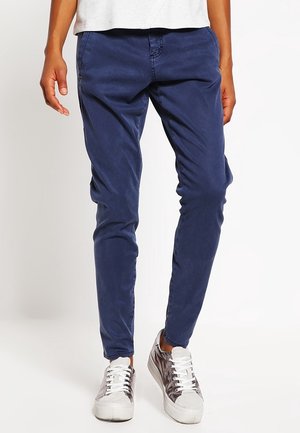 Person iført tætsiddende marineblå bukser og metalliske sølvfarvede sneakers, stående mod en ensfarvet lys baggrund.