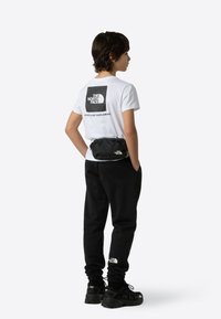 Tricou alb cu logo negru, borsetă neagr ă, pantaloni sport negri și adidași negri. Design simplu, croială casual și culori monocromatice.