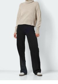 Personne portant un pull à col roulé beige, un pantalon noir ample et des baskets blanches, debout devant un fond clair uni.