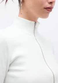 Λευκή μπλούζα με ψηλό λαιμό και ραβδώσεις, με ασημί φερμουάρ. Η κοντινή λήψη αναδεικνύει την υφή του υφάσματος και τη λεπτομέρεια του γιακά.