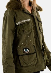 Chaqueta verde con ribete de piel, bolsillo decorado, detalles con tachuelas y diseño de ojo bordado. Presenta un ajuste casual y texturas variadas.