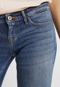 Jeans de denim azul ajustados con un acabado desgastado. Presentan un cierre delantero con botón, diseño de cinco bolsillos y acentos de costura en contraste.