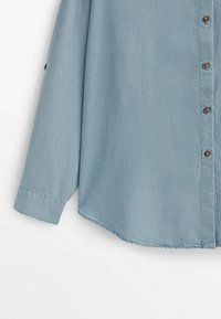 Chemise bleu clair à manches longues avec boutons marron et poignets cousus, vue du milieu du corps jusqu'à l'extrémité de la manche.